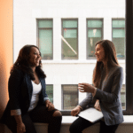 two women sit in a windowsill talking and smiling at work